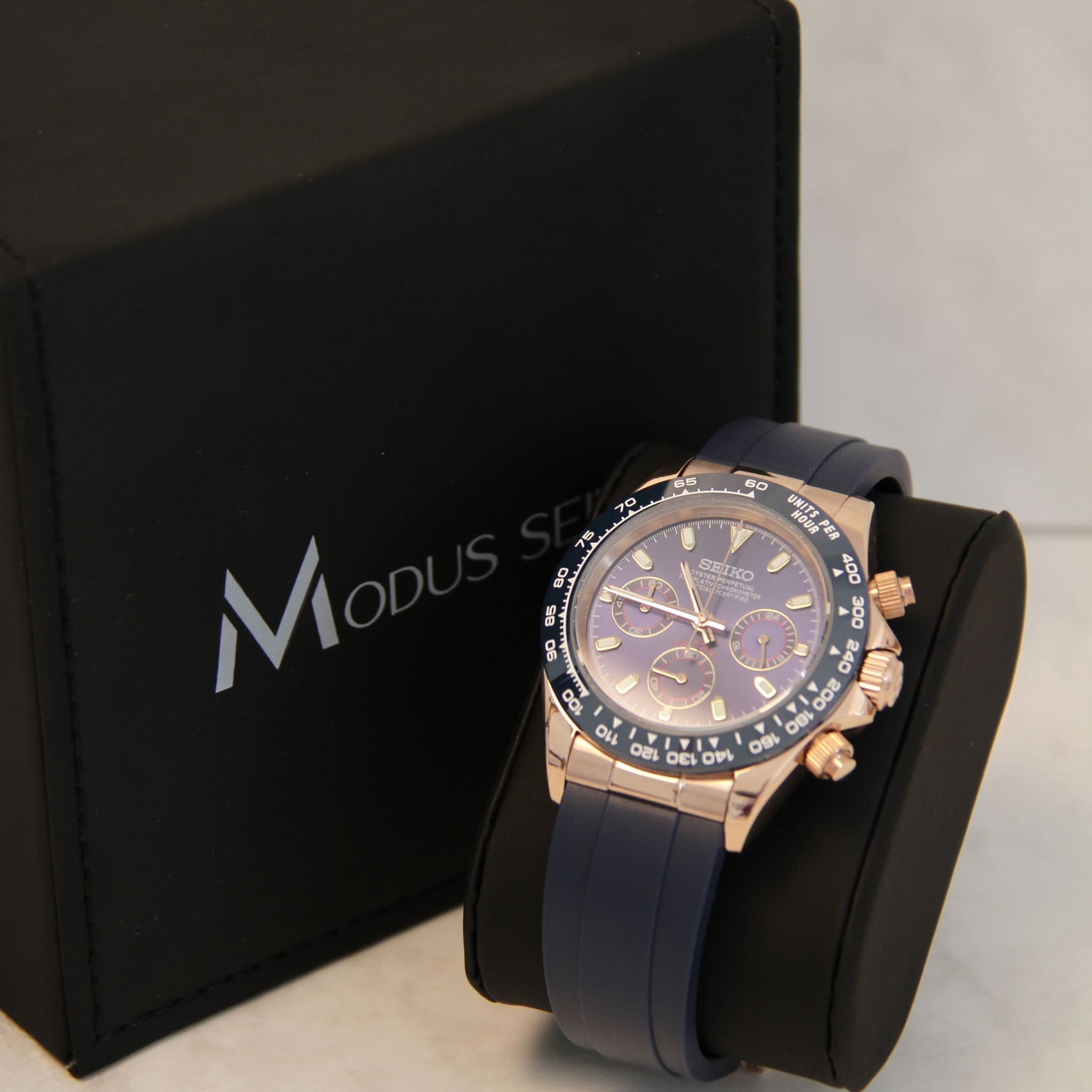 Montre Seiko Mod daytona or rose bleu sur coussin noir avec écrin en arrière plan.