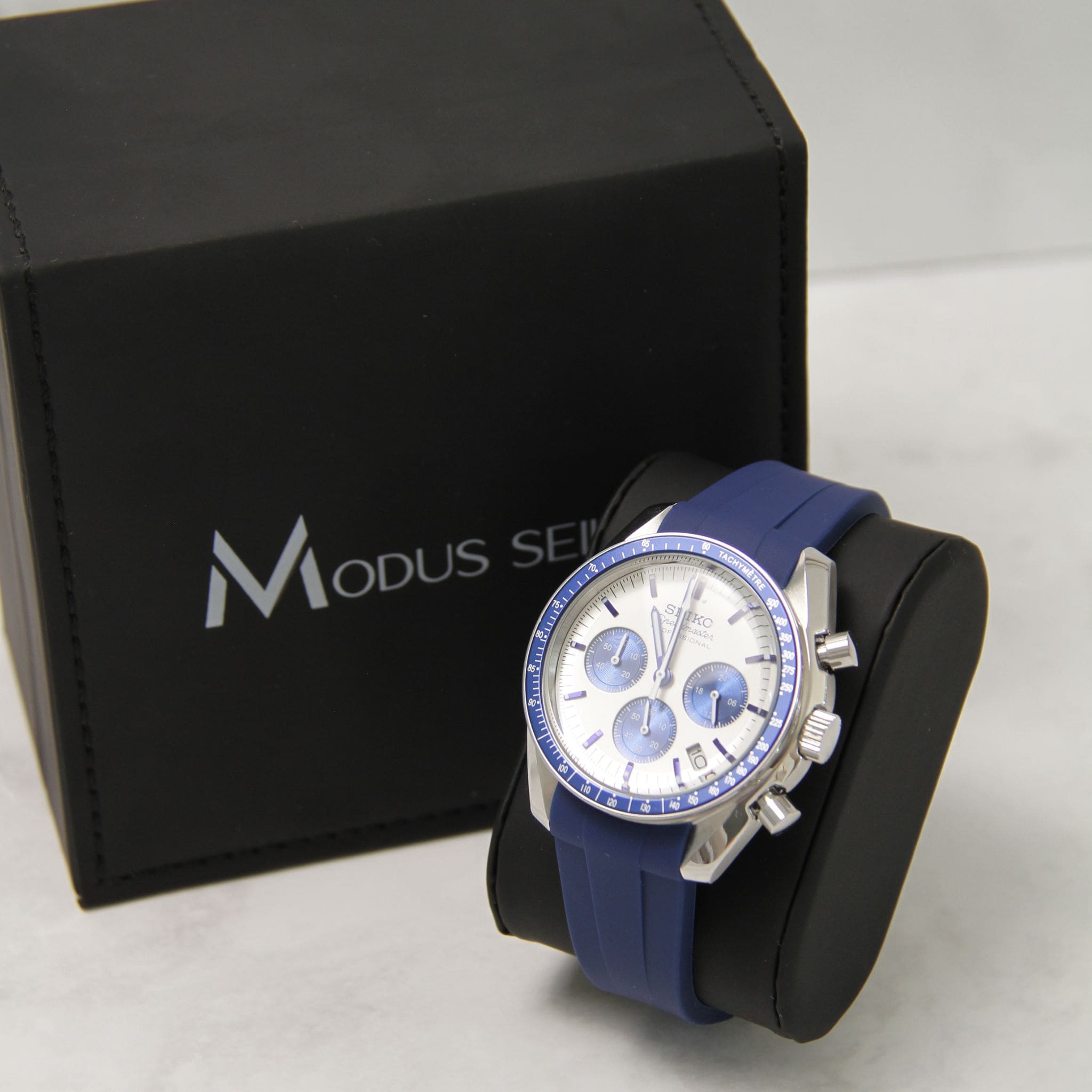 Montre Seiko Mod speedmaster quartz sur coussin noir avec écrin en arrière plan. 